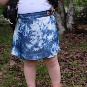 Op de afbeelding: Een jong persoon draagt een wit tanktop en een blauwe tie-dye denim rok met een bruine hartvormige knoop. Ze houden twee zwarte kuikens in hun armen en dragen veelkleurige sandalen.