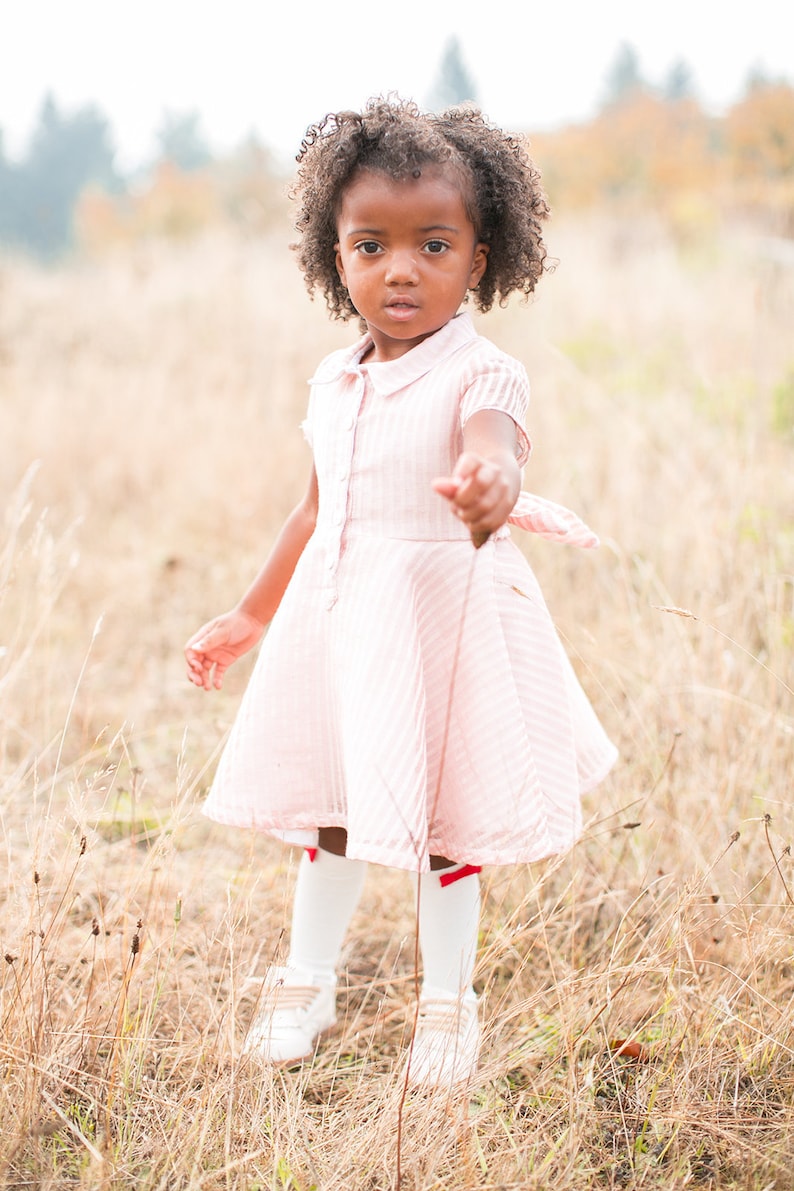 May include: A young girl with curly brown hair wearing a pink and white striped dress, white socks with red bows, and white shoes stands in a field of long grass.