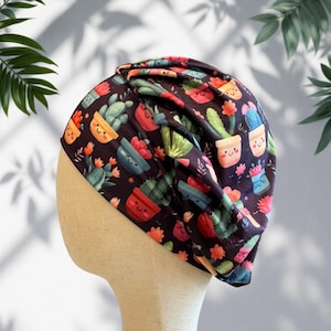 May include: A dark-colored fabric head wrap featuring a repeating pattern of various potted cacti with smiling faces. The head covering has a gathered, pleated design at the top and is displayed on a neutral-toned mannequin head form.