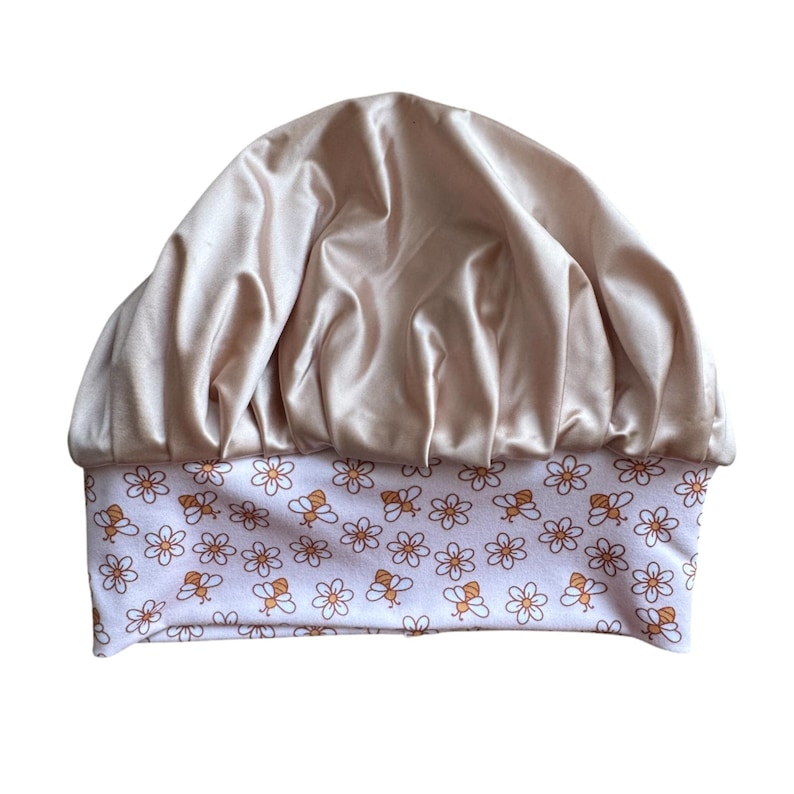 May include: A bonnet featuring a shiny, champagne-colored satin top section with gathered fabric folds. The wide, flat headband base is white and decorated with a repeating pattern of small orange-outlined daisies and brown-striped bumblebees.