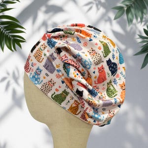 May include: A cream-coloured fabric turban hat featuring a repeating pattern of colourful cats in various poses, including orange, blue, green, and spotted designs, interspersed with small floral elements and paw prints, with gathered pleats at the front.