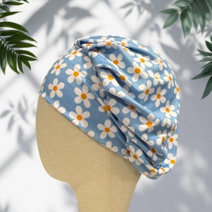 May include: A light blue fabric head covering featuring a repeating pattern of white daisies with yellow centers. The head covering has a gathered, pleated design on one side, creating a soft, draped appearance across the top and side of the form.