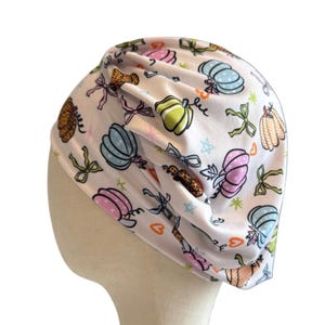 May include: A white fabric head covering featuring a repeating pattern of colorful pumpkins in shades of pink, blue, green, and orange, interspersed with small bows, stars, and heart shapes. The head covering has a gathered, pleated design.