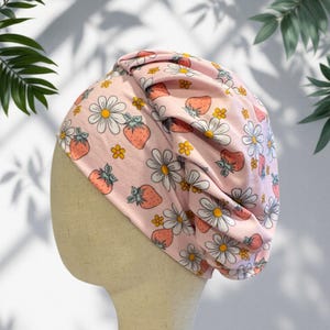 May include: A light pink fabric head covering featuring a repeating pattern of red strawberries, white daisies with yellow centers, and small yellow flowers. The head covering has a gathered, pleated design on one side and is displayed on a mannequin.
