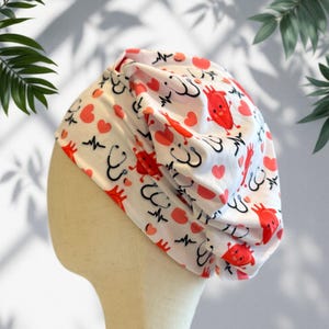 Puede incluir: Gorro quirúrgico blanco con un estampado repetido de corazones rojos, corazones de dibujos animados, estetoscopios y personajes en forma de corazón rojo. El gorro se muestra en una cabeza de maniquí.