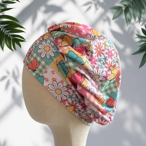 May include: A colorful turban featuring a patchwork design with floral and butterfly patterns. The fabric incorporates squares of pink, yellow, green, and white, creating a vibrant and eye-catching accessory. The turban is designed to be worn on the head.