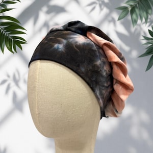May include: A tie-dye headband in shades of black, blue, and peach. The fabric is soft and gathered, creating a layered effect. The headband is displayed on a mannequin head, showcasing its shape and design. The accessory is suitable for various occasions.