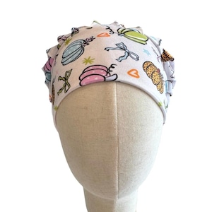 May include: A white scrub cap featuring a repeating pattern of colorful pumpkins, bows, and small orange hearts. The fabric has a gathered, pleated design around the crown, providing a structured fit. The print includes pastel pink, blue, and green pumpkin illustrations with various textures and outlines.