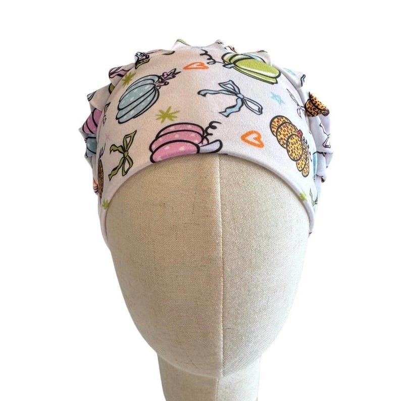 May include: A white scrub cap featuring a repeating pattern of colorful pumpkins, bows, and small orange hearts. The fabric has a gathered, pleated design around the crown, providing a structured fit. The print includes pastel pink, blue, and green pumpkin illustrations with various textures and outlines.