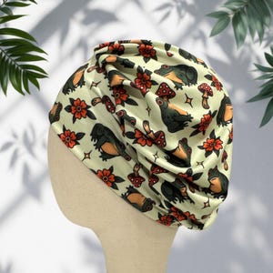 Puede incluir: Un turbante verde claro con un patrón repetido de ranas, setas y flores naranjas y marrones. El turbante está hecho de un material suave y sedoso y está diseñado para ser usado en la cabeza. El tema general es la naturaleza y la fantasía.