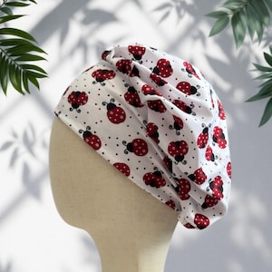 Puede incluir: Gorro de tela blanca con un estampado repetido de mariquitas rojas y puntos negros. Las mariquitas tienen antenas negras y están dispersas por la tela. El gorro se muestra en una cabeza de maniquí.
