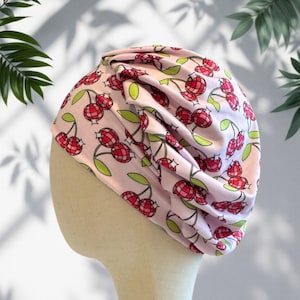 May include: A light pink turban-style head covering featuring a repeating pattern of red disco ball cherries with green leaves. The fabric is gathered at the top, creating soft folds and a rounded silhouette that fits over a mannequin head form.
