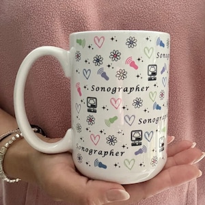 May include: White ceramic mug with a black and white design featuring the word "Sonographer" repeated multiple times. The design also includes hearts, flowers, and ultrasound symbols.