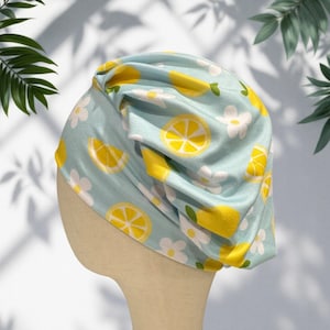 May include: A light blue turban-style head covering featuring a repeating pattern of bright yellow lemon slices and simple white flowers with yellow centers. The fabric is gathered at the top, creating soft folds and a textured, layered appearance.