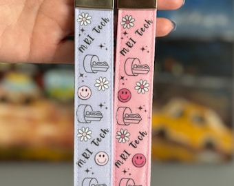 May include: Two keychains with a repeating pattern of an MRI machine, a smiley face, and a daisy. The text "MRI Tech" is repeated on each keychain. One keychain is pink and the other is lavender.