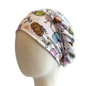 May include: A white slouchy beanie featuring a repeating pattern of pumpkins in various colors including leopard print, light blue, and green, interspersed with small pink bows, orange hearts, and tiny star shapes across the fabric surface.