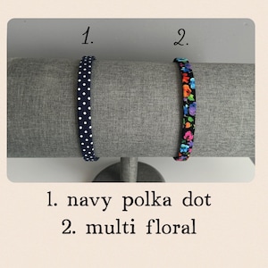 Puede incluir: Dos diademas en un expositor. La diadema de la izquierda es azul marino con lunares blancos. La diadema de la derecha es negra con un estampado floral multicolor. El texto "1. navy polka dot" y "2. multi floral" est&aacute; debajo de las diademas.