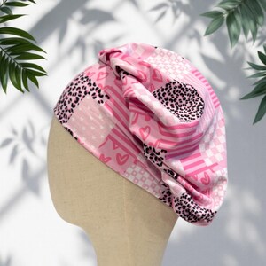May include: A pink turban-style head wrap with a patchwork design. The pattern includes hearts, leopard print, stripes, and checkered squares. The head wrap is displayed on a mannequin head.