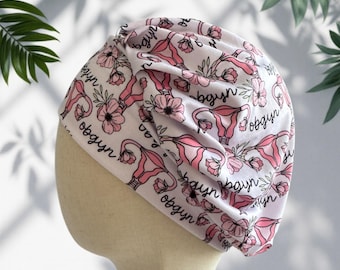 May include: A white scrub cap featuring a repeating pattern of pink anatomical uterus illustrations, pink flowers, and the black cursive text "obgyn". The cap has a gathered, pleated design at the front and is displayed on a neutral mannequin head.