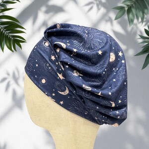 May include: A dark blue turban-style head covering with a celestial design. The fabric features white and light pink illustrations of the sun, moon, stars, and clouds. The head covering is displayed on a mannequin head.