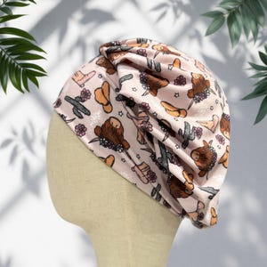 May include: A light pink turban-style head covering featuring a repeating pattern of brown highland cows with floral crowns, green cacti, and tan cowboy boots. The fabric is gathered at the top, creating soft folds and a textured appearance.
