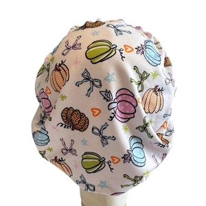 May include: A white scrub cap featuring a repeating pattern of colorful pumpkins in shades of pink, orange, green, and blue, interspersed with small bows, orange hearts, and tiny stars. The fabric has a gathered design with a rounded top shape.