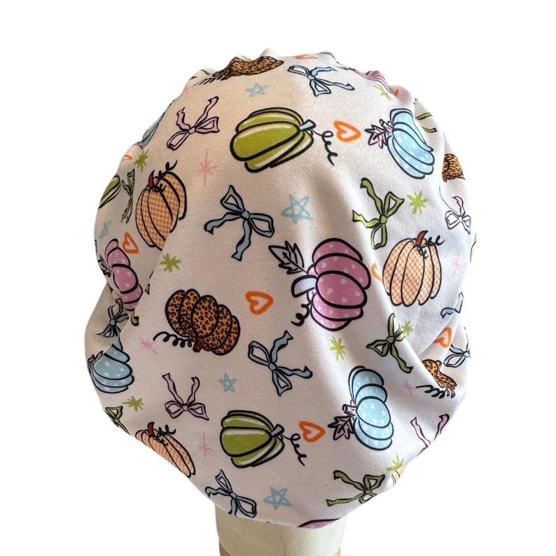 May include: A white scrub cap featuring a repeating pattern of colorful pumpkins in shades of pink, orange, green, and blue, interspersed with small bows, orange hearts, and tiny stars. The fabric has a gathered design with a rounded top shape.