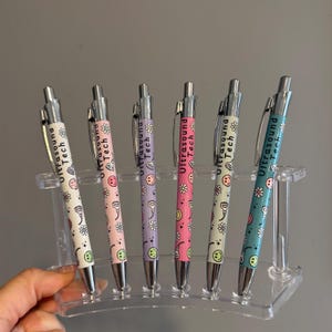 Może przedstawiać: Sześć długopisów ze srebrnymi skuwkami i kolorowymi korpusami. Każdy długopis ma białe tło z powtarzającym się wzorem kwiatów i uśmiechniętych twarzy. Tekst "Ultrasound Tech" jest nadrukowany na każdym długopisie czarnym kolorem.