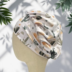 May include: A cream-colored turban hat featuring a repeating pattern of green cacti and abstract orange shapes. The fabric has a gathered, pleated design that creates soft folds across the surface, providing a textured look to the headwear.