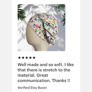 May include: A white fabric head covering featuring a colorful pattern of pink flowers, green leaves, and small insects. The head covering is displayed on a neutral-toned mannequin head, showcasing a gathered, pleated design with a soft, flexible fit.