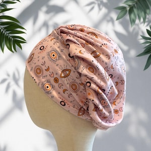 May include: A light pink turban-style head covering featuring a repeating pattern of orange and black stylized eyes, crescent moons, and circular sun motifs. The fabric is gathered at the side to create soft folds and a draped appearance.