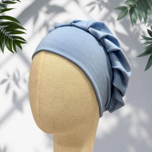 May include: A light blue jersey fabric turban hat featuring a smooth, fitted crown and a wide, pleated band that wraps around the back and sides, creating a soft, gathered texture. The head covering is displayed on a beige mannequin head.