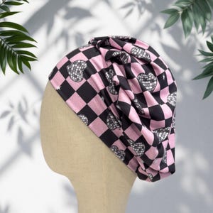 May include: A pink and black checkered turban-style hat with disco ball heart patterns. The hat is made of soft fabric and is displayed on a mannequin head. The design features a repeating pattern of black squares, pink squares, and disco ball hearts.