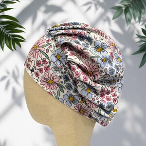 Puede incluir: Un turbante con estampado floral en tonos pastel de rosa, azul y amarillo sobre fondo blanco. El turbante, hecho de tela suave, está diseñado para ser usado en la cabeza. El diseño floral presenta margaritas y otras flores pequeñas.