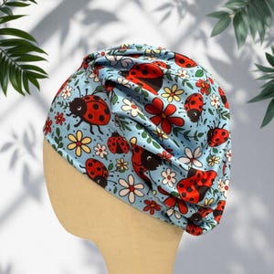 May include: A light blue turban-style head covering featuring a repeating pattern of red ladybugs with black spots, interspersed with small white and yellow flowers and green leaves. The fabric is gathered at the top to create a pleated effect.