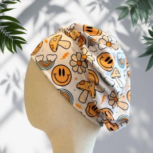 May include: White beanie hat with a repeating pattern of orange smiley faces, mushrooms, flowers, rainbows, and lightning bolts. The hat is displayed on a mannequin head. The design has a retro, groovy aesthetic.