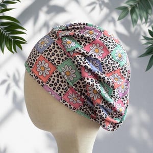May include: A gathered beanie hat featuring a repeating patchwork pattern of white daisies with yellow centers on pastel pink, blue, and green squares, interspersed with sections of a black and tan leopard print design on a light background.