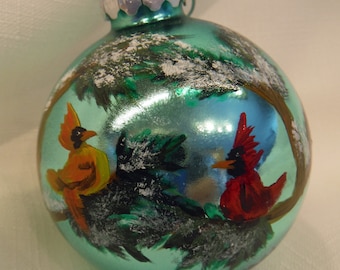 Puede incluir: Adorno navideño de cristal color turquesa con una escena invernal pintada a mano. Dos cardenales, uno naranja y otro rojo, se posan en una rama. El adorno tiene una tapa de metal plateado y un lazo para colgar. El fondo presenta un bosque nevado.