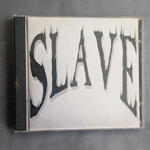 May include: A CD case with the word "SLAVE" in large, stylized, dark gray letters against a light gray background. The CD case is in a clear plastic sleeve, and the spine is black.