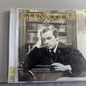 以下が含まれることがあります： CDケース。金色の文字で「GLENN GOULD ...And Serenity」と書かれています。CDの画像は、スーツを着て座って考え込んでいる男性の白黒写真です。背景には本棚があります。