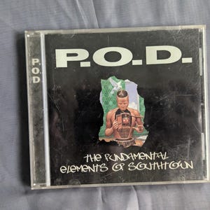 May include: A CD case featuring the band name "P.O.D." in white block letters against a black background. The album title "The Fundamental Elements of Southtown" is below the band name. The cover art includes a figure and a green and white design.