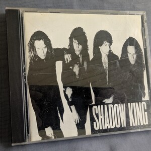 Può includere: Un'immagine in bianco e nero di una rock band su una custodia per CD. I membri della band indossano abiti scuri, con i capelli lunghi. Il nome della band, "SHADOW KING", è stampato in lettere bianche in basso sulla copertina.