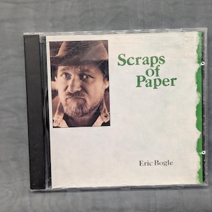 May include: A compact disc case featuring a white cover with a green torn-paper edge. The cover displays a sepia-toned portrait of a bearded person wearing a hat, with the green text Scraps of Paper and the black text Eric Bogle printed on it.