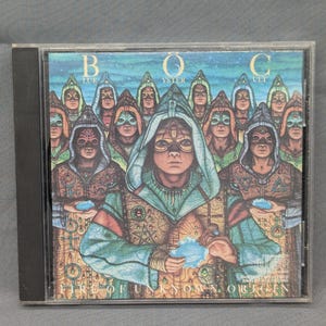 May include: A compact disc case featuring the album cover for "Fire of Unknown Origin" by BÖC. The artwork depicts a group of hooded figures in teal and brown tones, holding blue orbs. The album title is at the top.