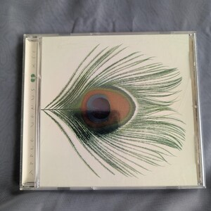 May include: A CD case featuring the album "Apple Venus Volume 1" by XTC. The cover art displays a detailed illustration of a peacock feather eye in shades of green, brown, and blue against a white background. The CD case is clear plastic.