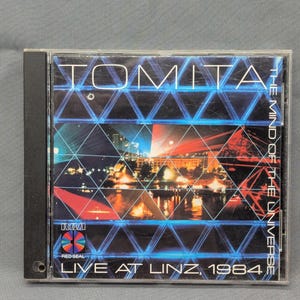 May include: A CD case featuring the album "Tomita - The Mind of the Universe: Live at Linz, 1984." The cover has a geometric design with a cityscape and the RCA Red Seal logo. The text is in white and blue.