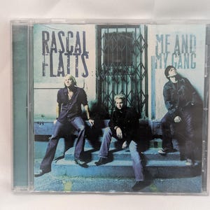May include: A Rascal Flatts Me and My Gang music CD in a clear plastic jewel case. The cover art features three individuals sitting on blue concrete steps in front of a metal security gate, with the band name and album title in black text.