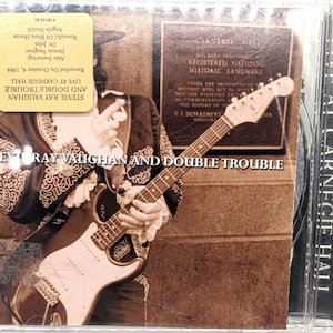 May include: CD case for "Stevie Ray Vaughan and Double Trouble: Live at Carnegie Hall." The cover features a sepia-toned image of a musician playing a guitar, with text overlay. The CD case is in a clear plastic sleeve.