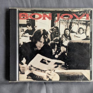 May include: A Bon Jovi "Cross Road" CD case. The album cover features a black and white photo of the band members. The text "BON JOVI" is in red, with "CROSS ROAD" in white. The case is in a clear plastic sleeve.