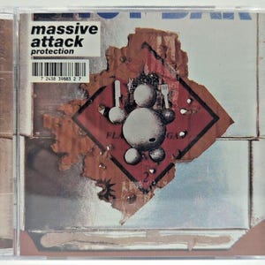 May include: A CD case featuring the album "Protection" by Massive Attack. The cover art includes a distressed design with a cartoon figure holding a knife and fork, set against a red diamond hazard sign. The album title and artist name are visible.